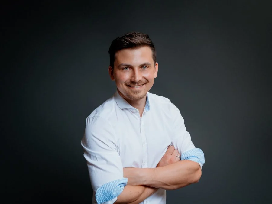 Lächelndes Teammitglied der Steuerkanzlei Patrick Herzer mit verschränkten Armen vor dunkelgrauem Hintergrund